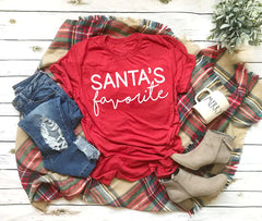 Red t-shirt with 'Santa's favorite' text on a plaid blanket with denim shorts, boots, and a mug.