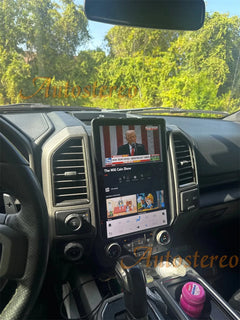 Car dashboard with a mounted tablet displaying a news app, surrounded by trees outside.